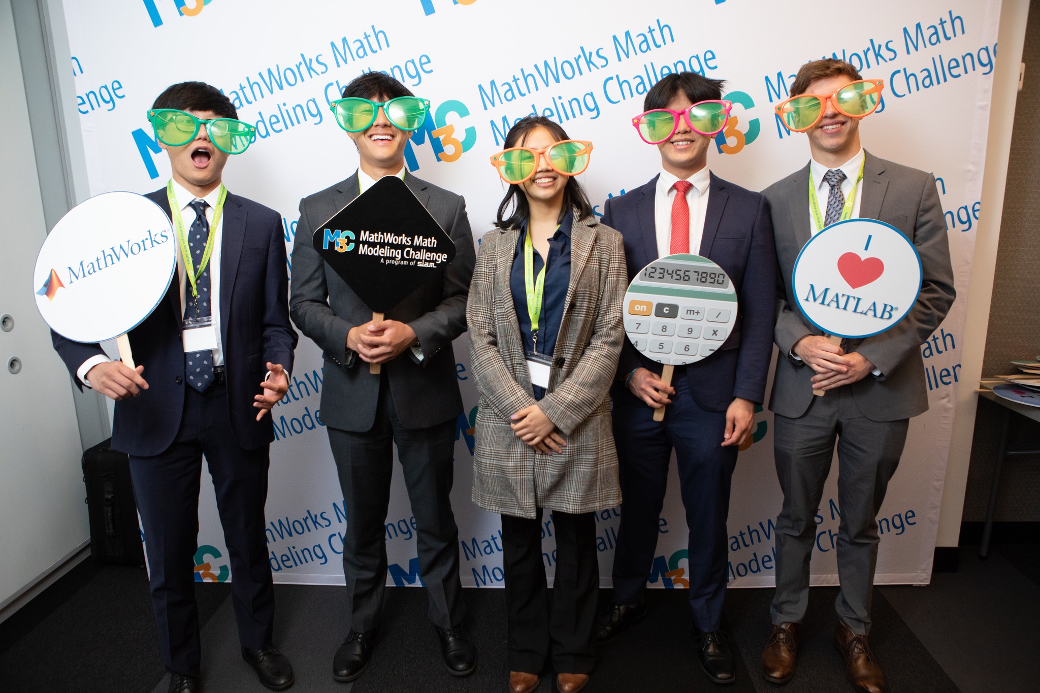 Five students posing in front of a photo wall wearing silly glasses and holding MathWorks props