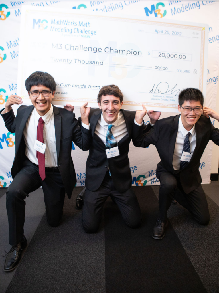 Three students kneeling and holding a giant check on their shoulders.